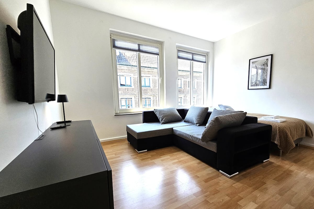 Un salon moderne avec un canapé d’angle, un téléviseur à écran plat et une grande fenêtre équipée de stores.