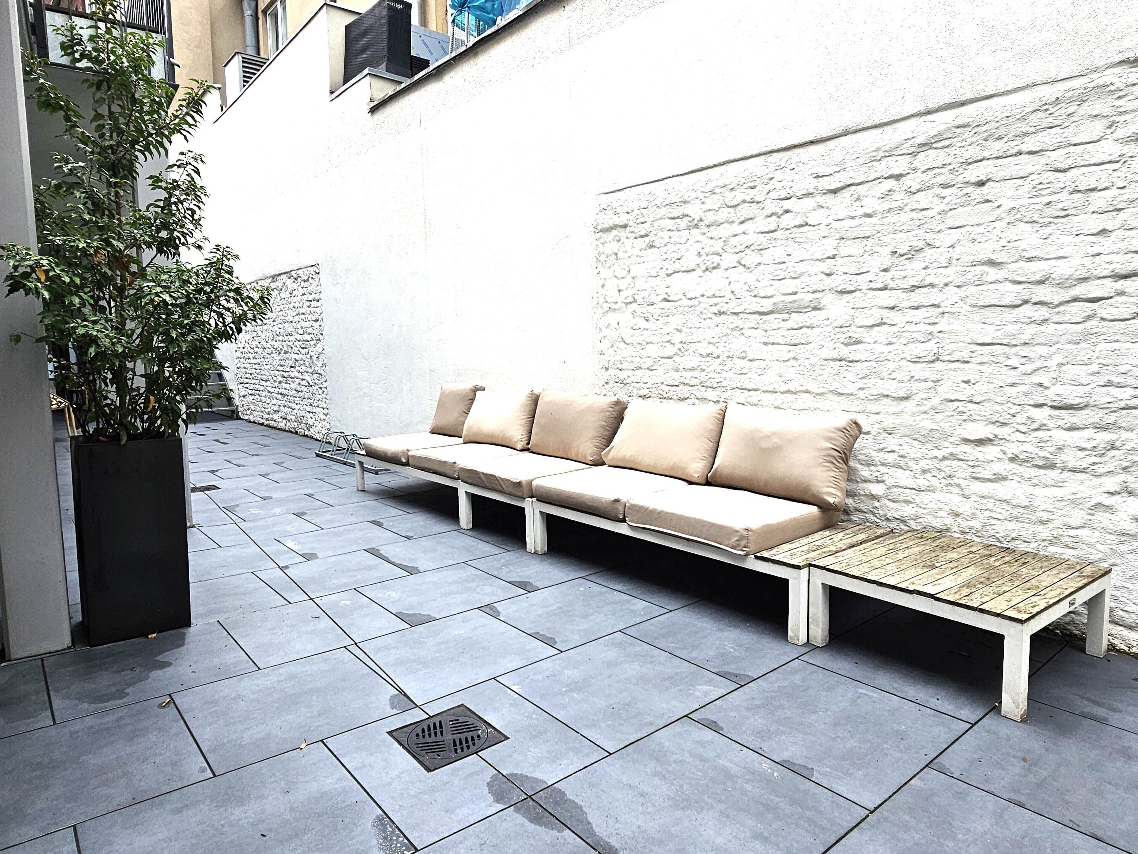 Een modern buitgedeelte met een beige bank tegen een witte, gestructureerde muur, een potplant aan de linkerkant en een houten bankje aan de rechterkant.