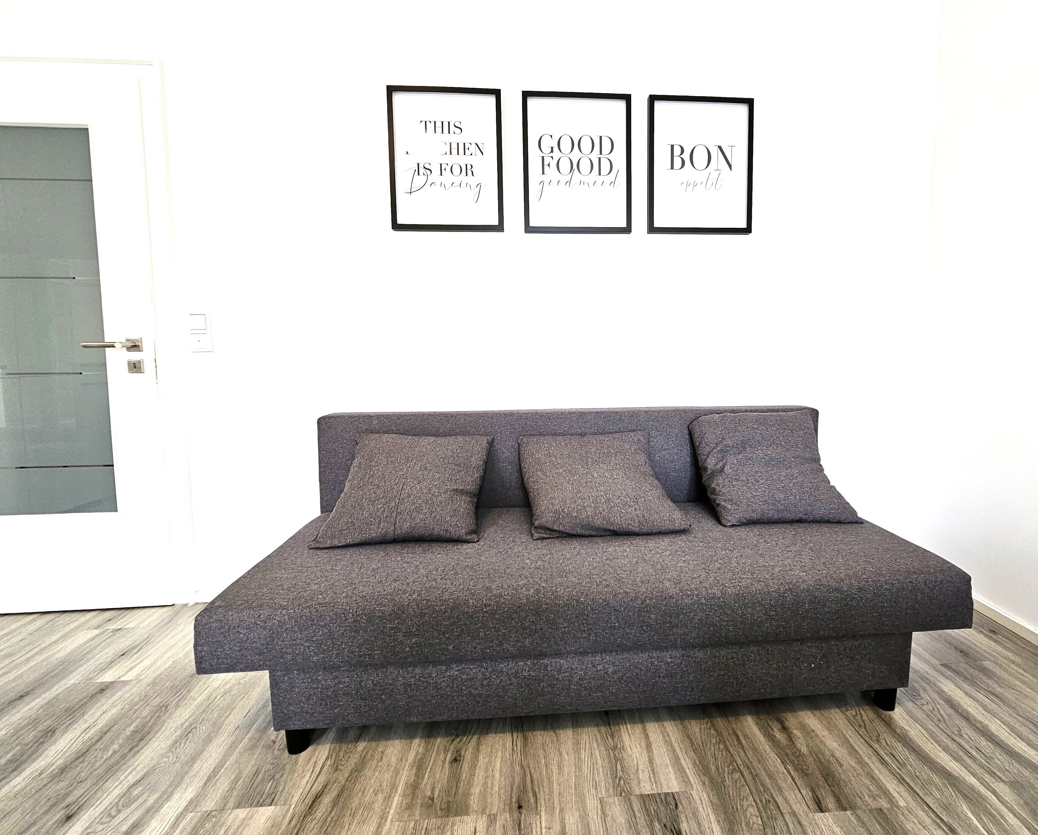 A grey sofa with three cushions in a room with a wooden floor and a white door. Three framed signs are hung on the wall above the sofa.