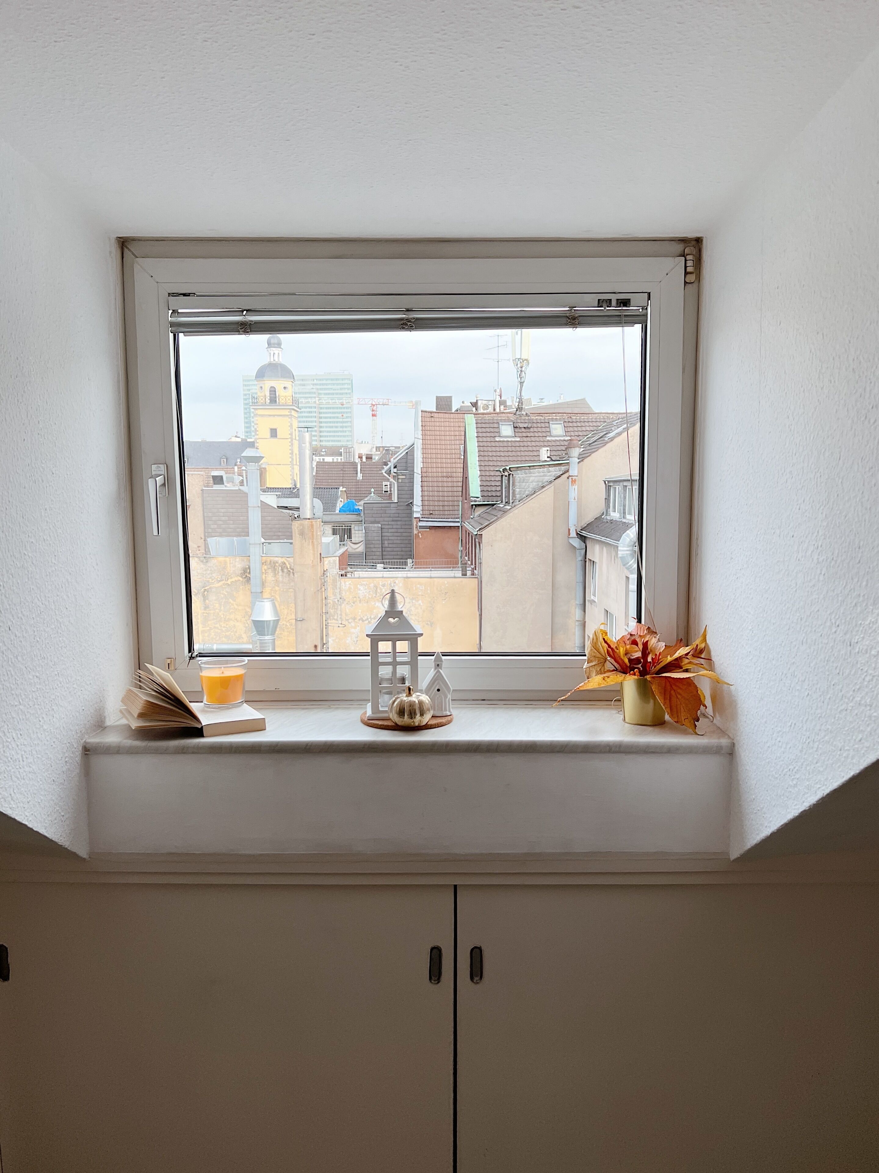 Ein Fenster mit Blick auf eine Stadtlandschaft, ein Buch, eine Kerze, ein Laternenlicht und eine Vase mit Blumen auf einem Regal.