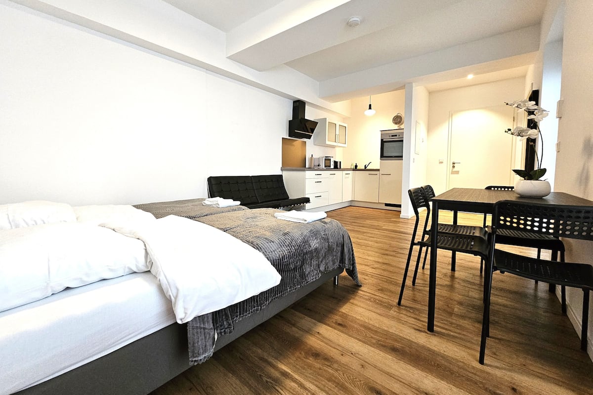 Une chambre moderne avec un lit, un petit bureau et une chaise. On aperçoit à l’arrière-plan un coin cuisine avec des placards et des appareils électroménagers.