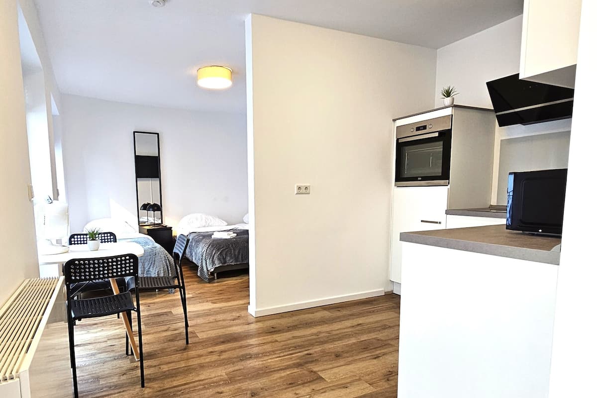 Une cuisine moderne équipée d’un four à micro-ondes, d’un four traditionnel et d’un petit plan de travail. On y trouve également un coin repas avec une table et des chaises. La pièce est dotée d’un parquet et de murs blancs.