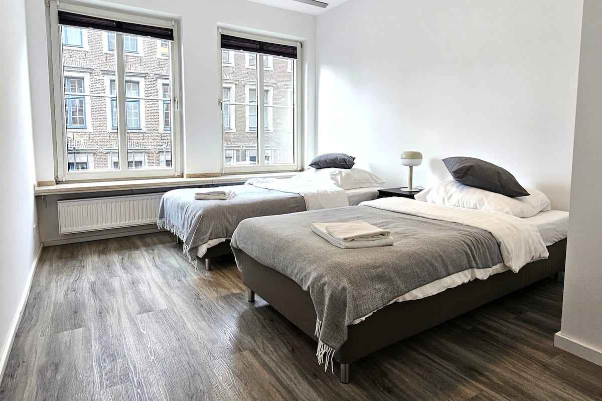 Une chambre moderne avec deux lits, un radiateur et une vue sur les bâtiments, visible à travers de grandes fenêtres.