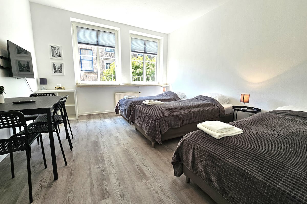 Une chambre avec deux lits, un bureau et une chaise. Elle est équipée de fenêtres avec stores, d’une lampe et d’un téléviseur fixé au mur.