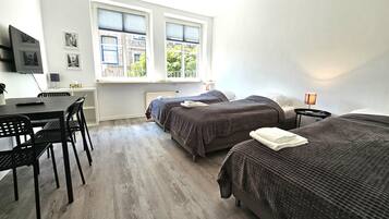 Ein Zimmer mit zwei Betten, einem Schreibtisch und einem Stuhl. Es gibt Fenster mit Jalousien, eine Lampe und einen an der Wand befestigten Fernseher.