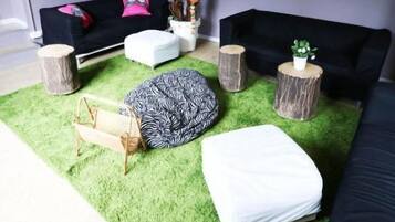 Une pièce dotée d’un sol recouvert d’un gazon synthétique vert, de deux canapés noirs, d’un pouf blanc, d’un fauteuil en forme de poire et d’un grand palmier artificiel.