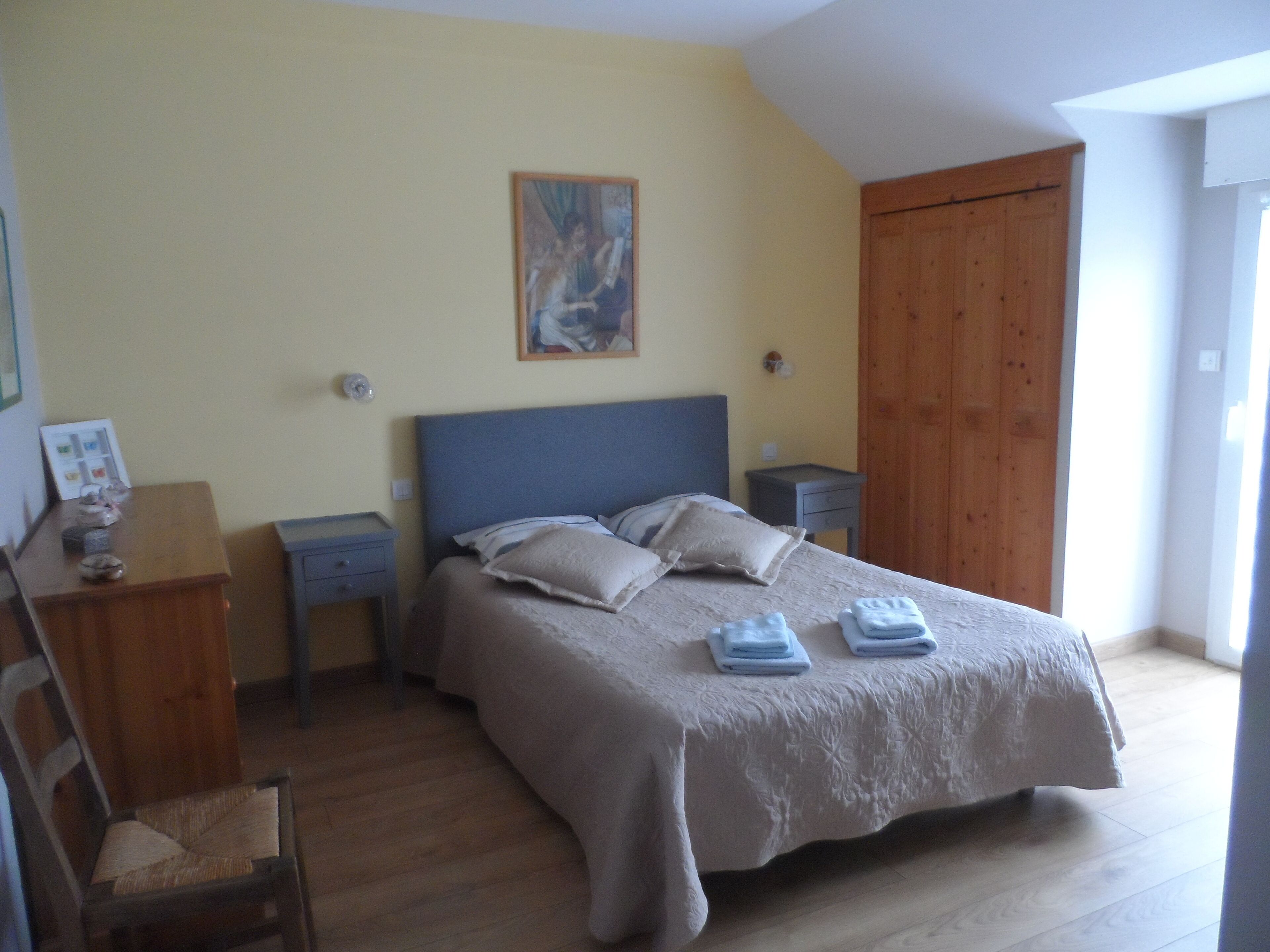 A bedroom with a large bed, two bedside tables, a wooden wardrobe, a chair, and a painting on the wall.