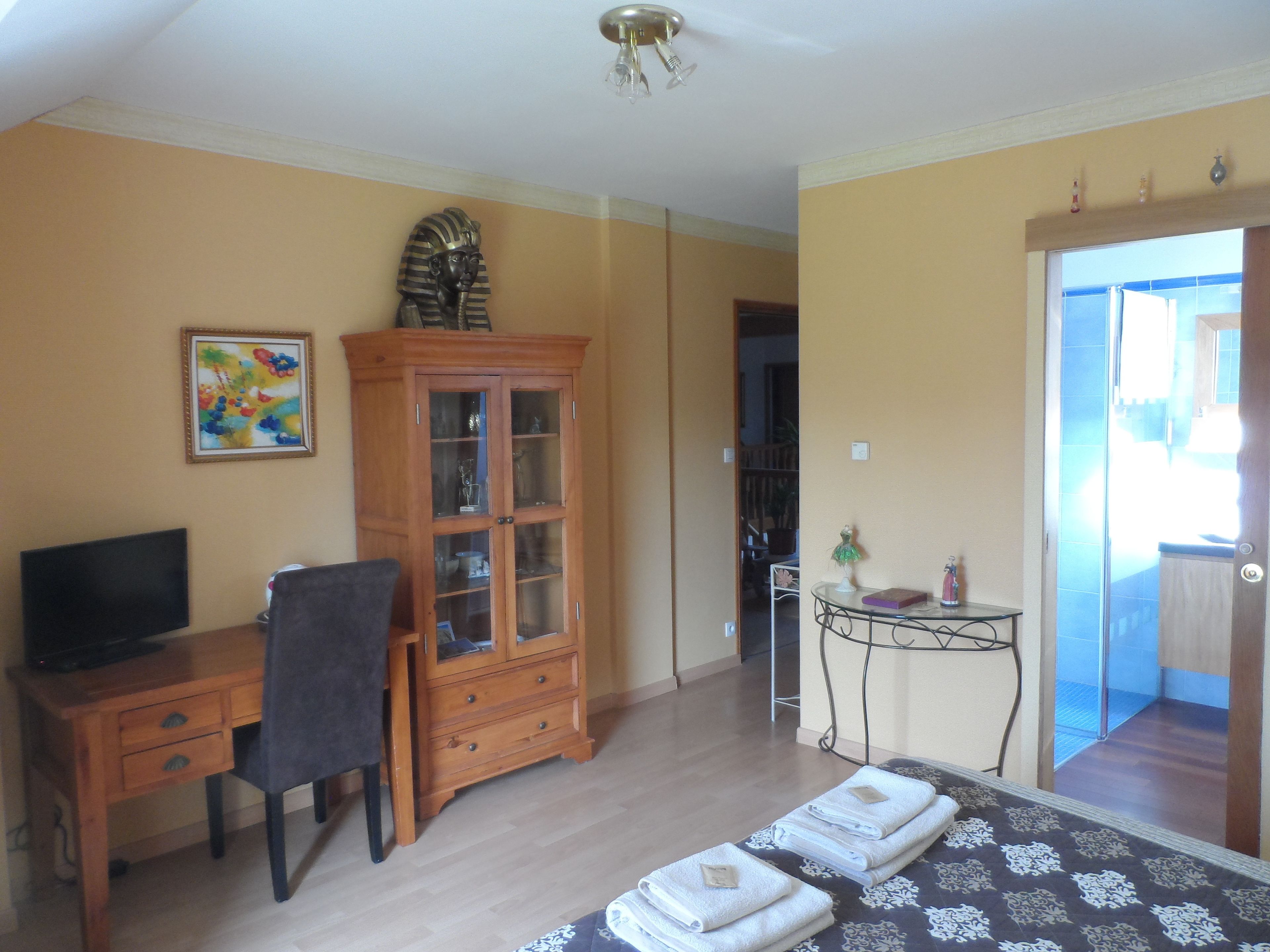 A bedroom with a bed, a desk with a chair, a cabinet, a painting, and a small table with a lamp.