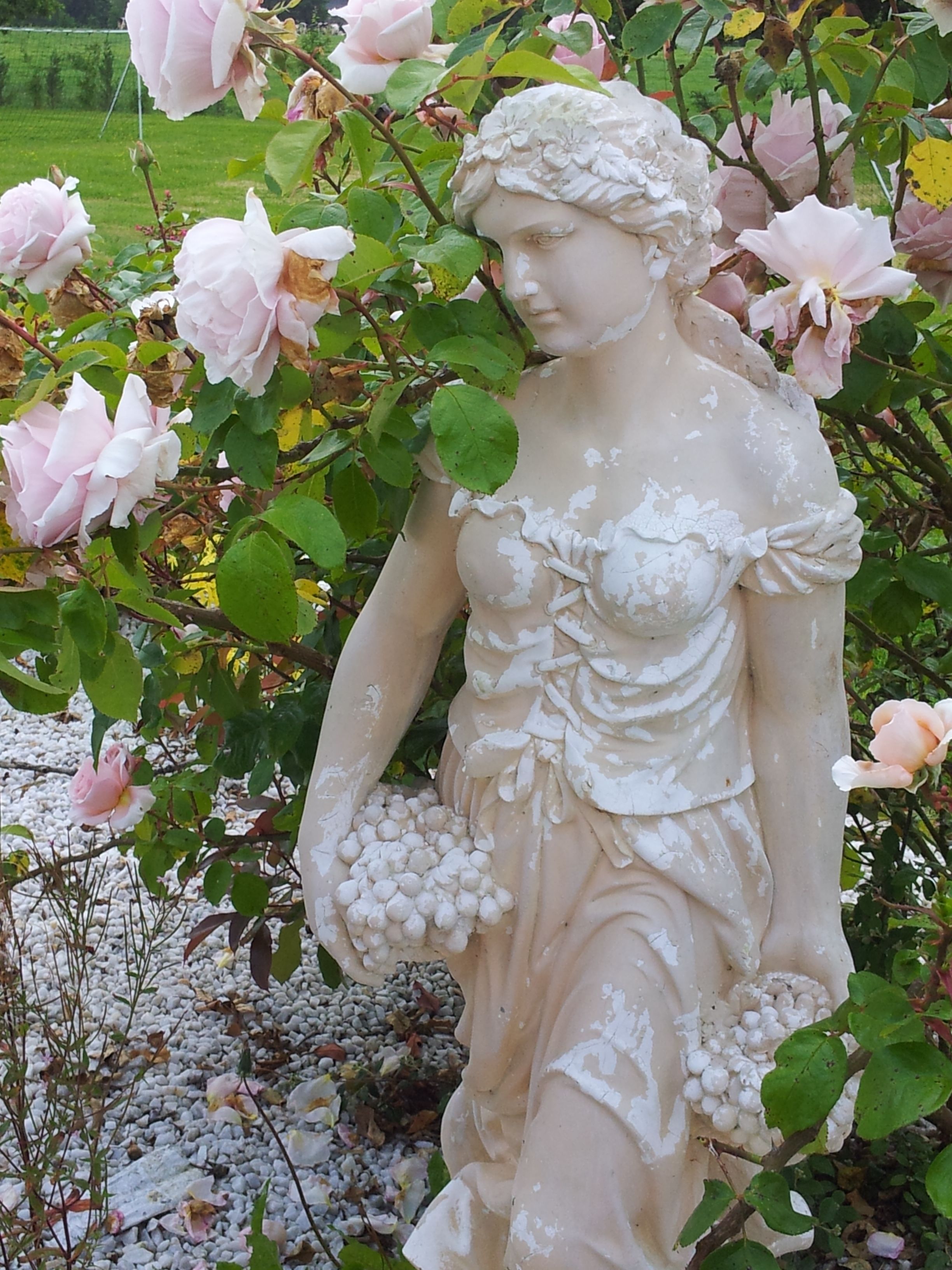 A statue of a woman holding grapes, surrounded by pink roses and greenery.
