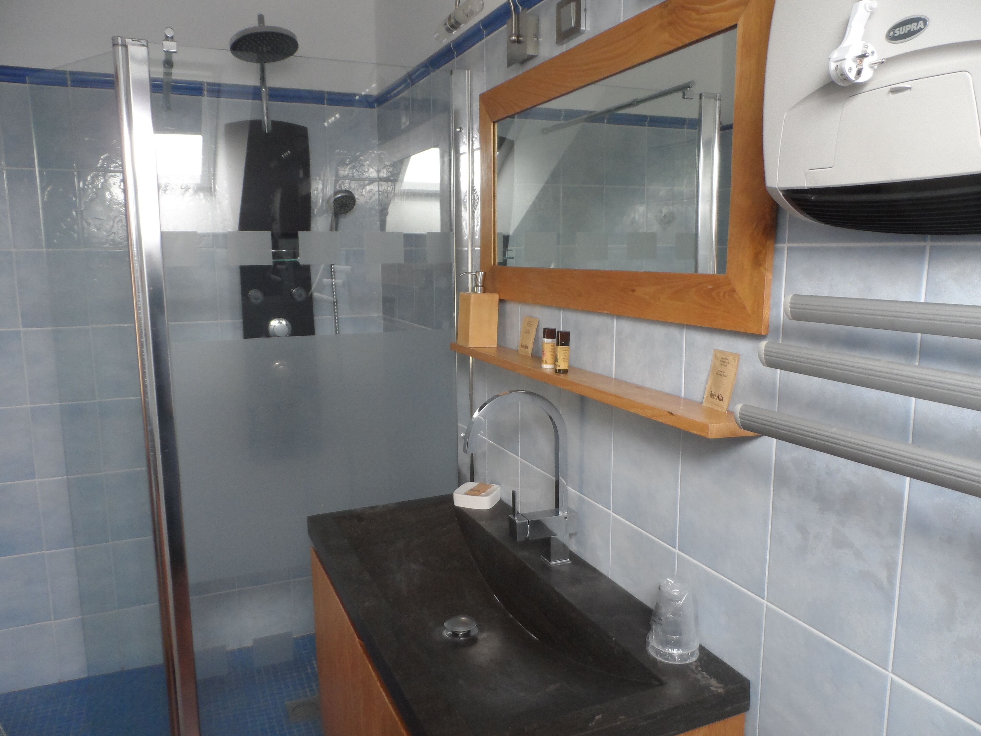 Un baño con cabina de ducha de vidrio, un lavabo negro, un estante de madera con artículos de aseo y un espejo sobre el lavabo.