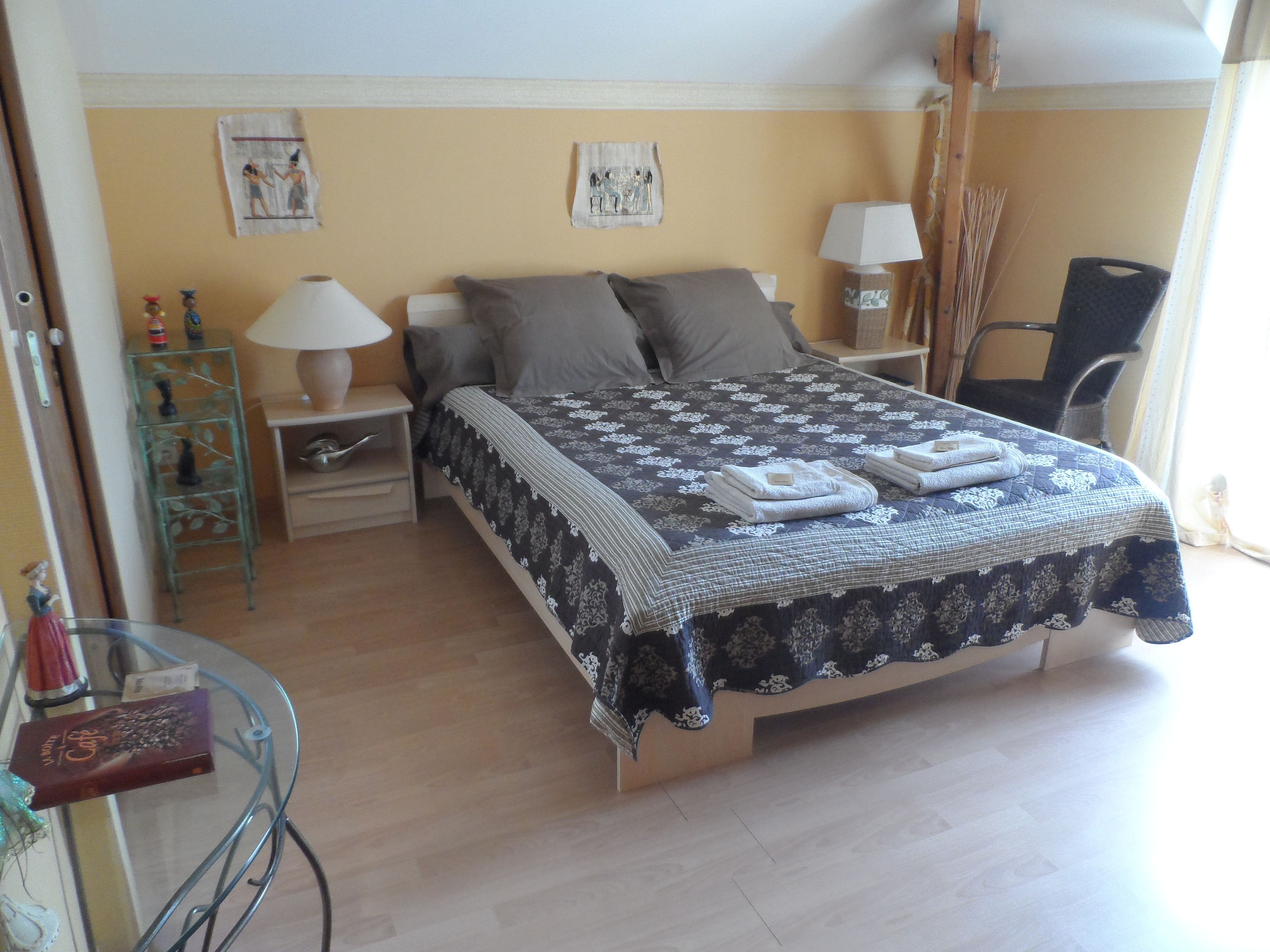 Una cama bien hecha con una colcha estampada y almohadas, una mesita de noche con lámpara, una silla y un pequeño estante con adornos.