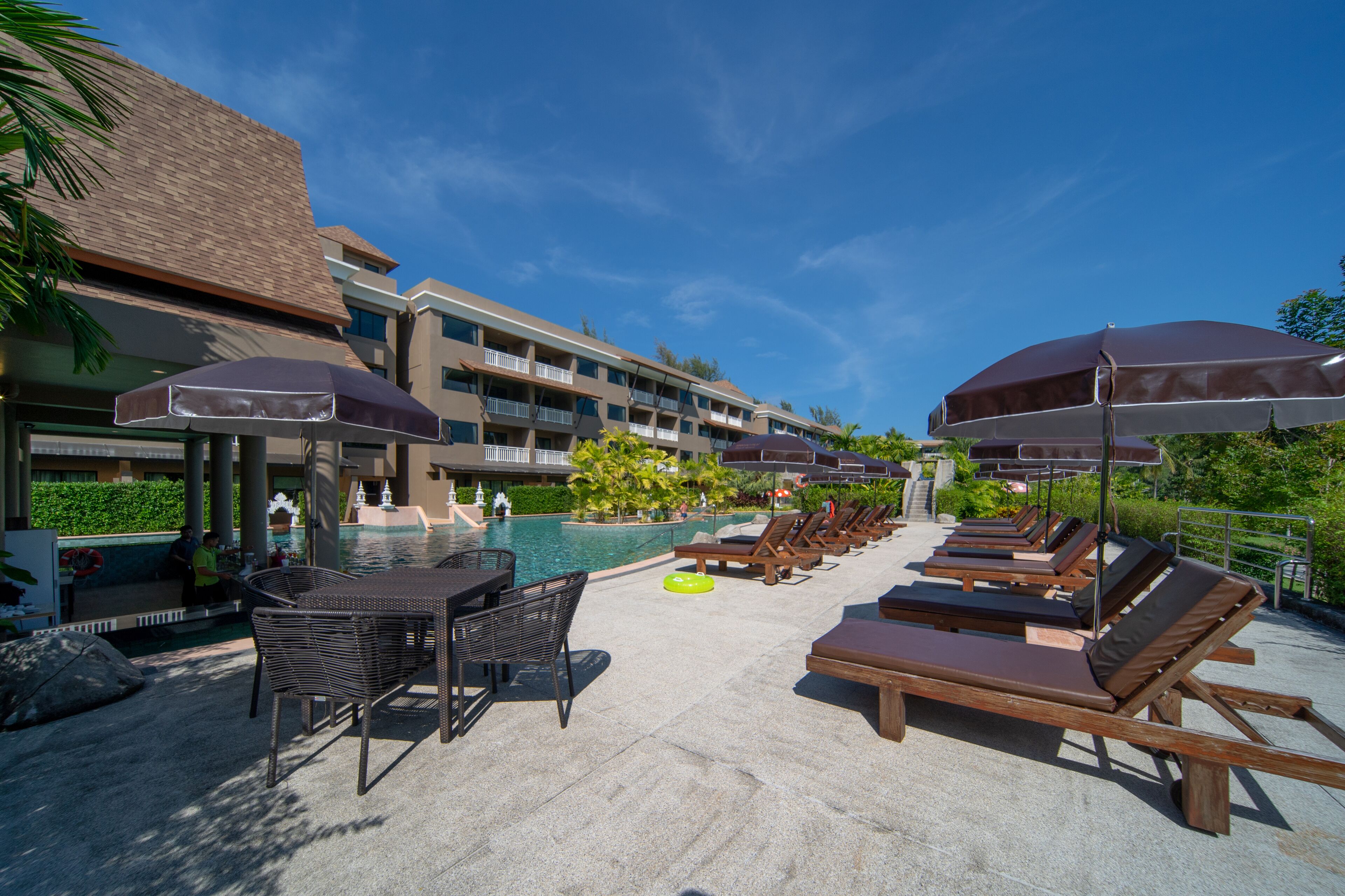 Zona d'una piscina de resort amb gandules i ombrel·les, piscina i un edifici amb balcons.
