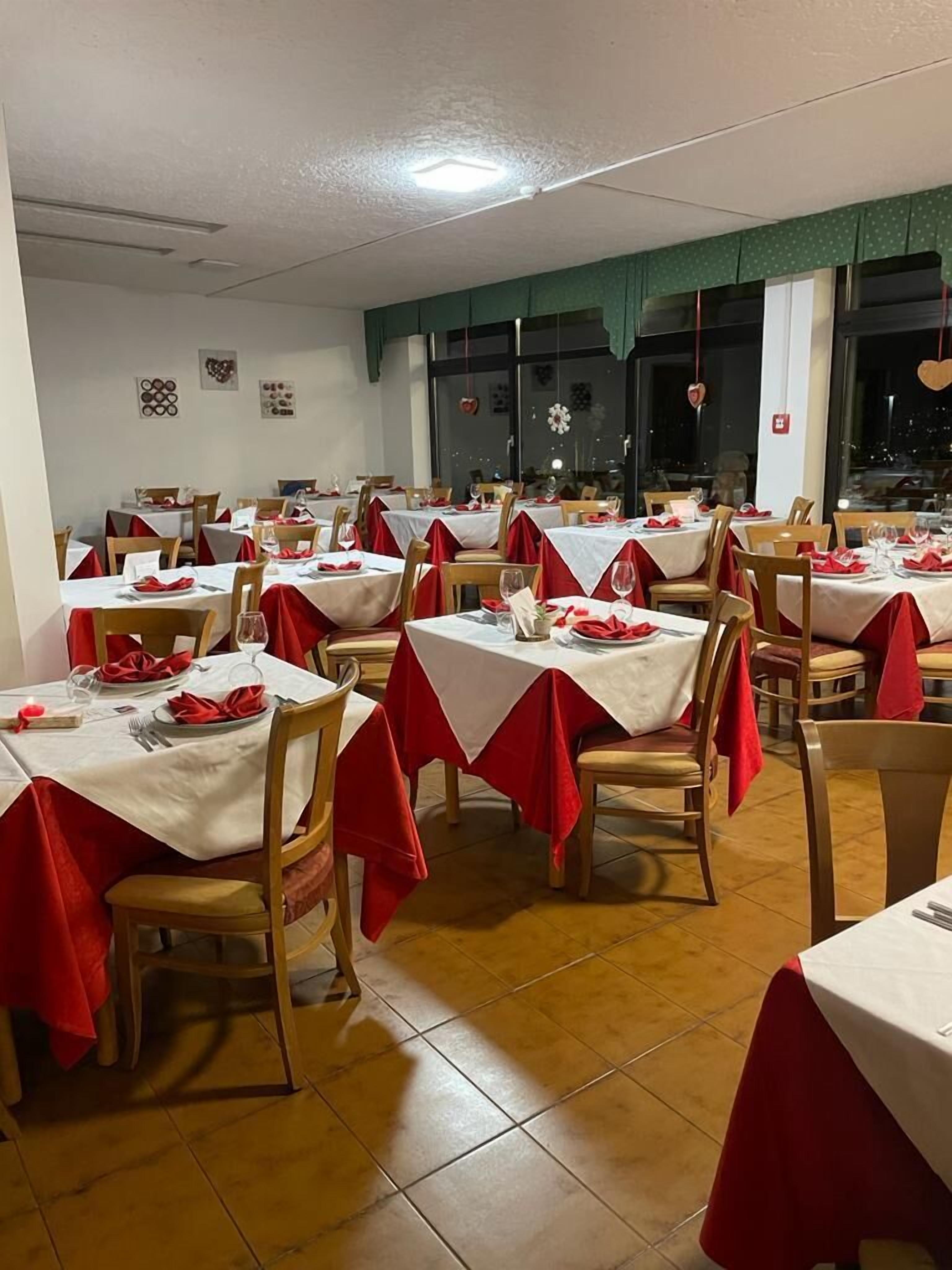 En restaurang med flera bord, vart och ett täckt med ett vitt bordsduks och röda bordstabletter. Rummet har trästolar och ett klinkergolv.