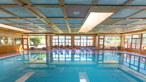 An indoor swimming pool with clear blue water, wooden beams on the ceiling, and several lounge chairs around the pool. - Hotel Sancamillo (Dimaro Folgarida)