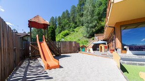 A backyard with a wooden fence, two orange slides, a sandy play area, and a small building with a red roof. - Hotel Sancamillo (Dimaro Folgarida)