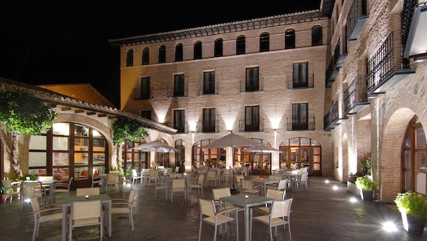 An outdoor cafe area with tables and chairs, surrounded by brick buildings with large windows and balconies. - Hotel Cienbalcones (Daroca)