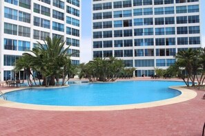 A swimming pool surrounded by palm trees and a modern building with multiple floors and large windows.