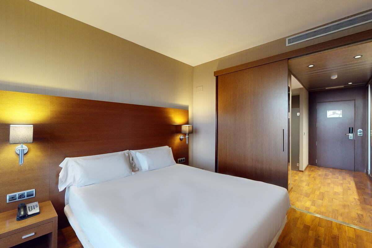A hotel room with a large bed, wooden headboard, two bedside lamps, a nightstand with a phone and a lamp, and a door leading to a bathroom.