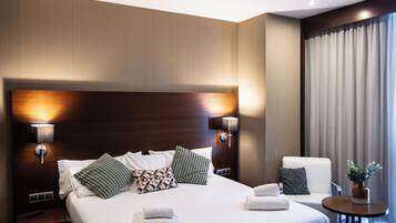 Een hotelkamer met een groot bed, twee bedlampen, een stoel, een klein tafeltje en een raam met gordijnen.