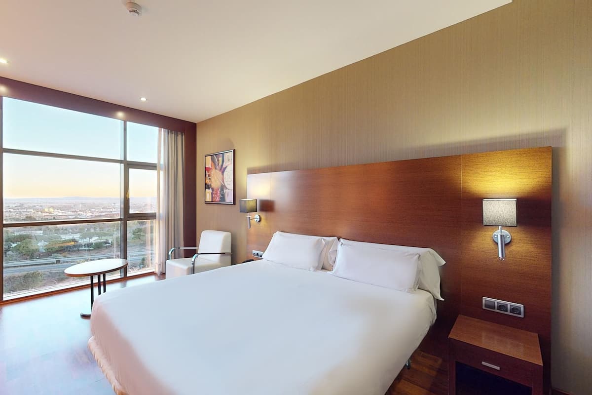 A modern hotel room with a large bed, a chair, a nightstand, and a view of the city through large windows.