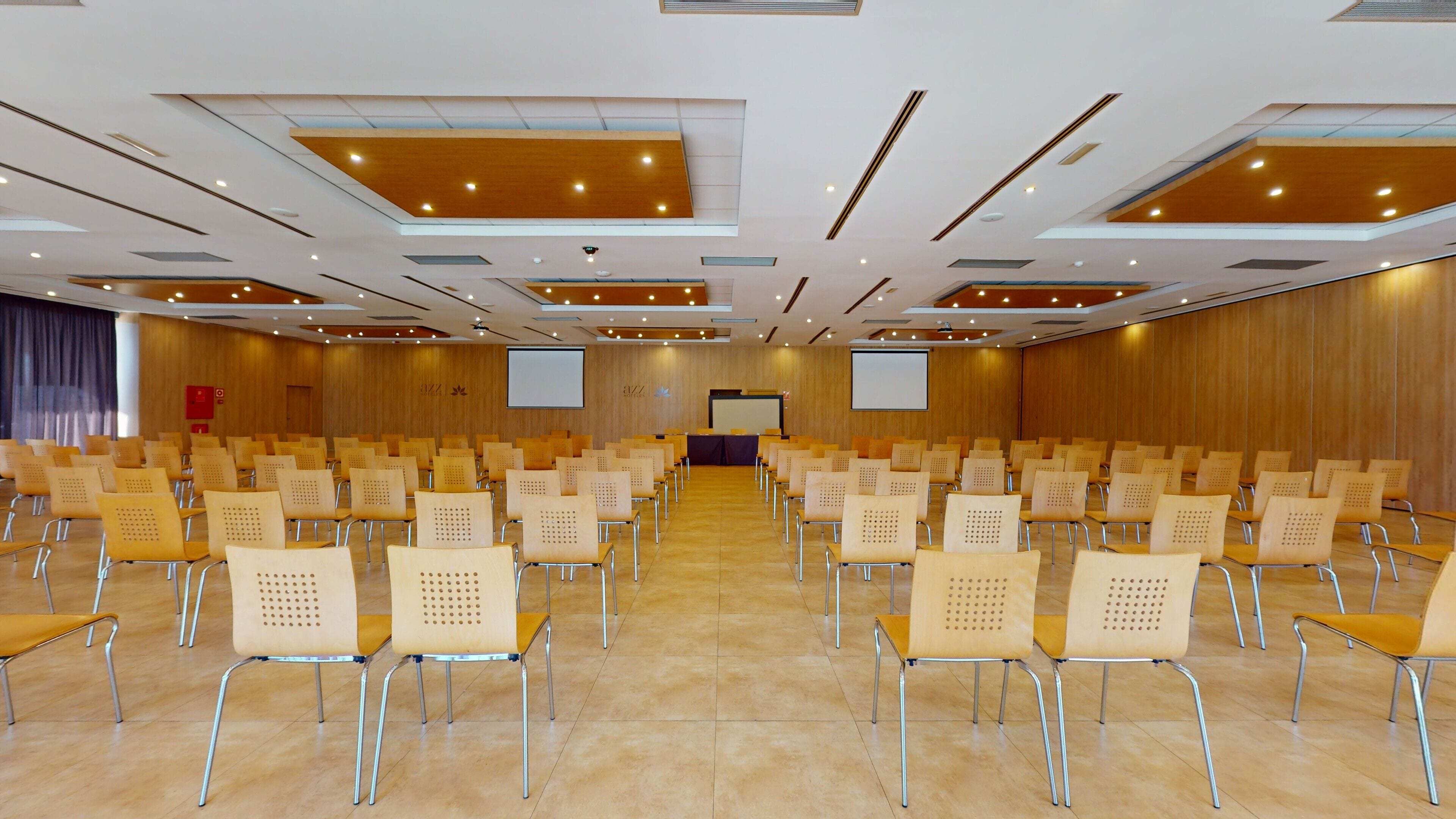 A conference room with rows of chairs, a stage with a projector screen, and a wooden panel wall.