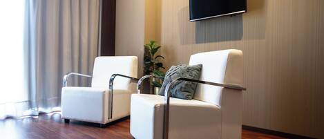A modern hotel room with two white leather armchairs, a flat-screen TV mounted on the wall, and a large window with curtains.