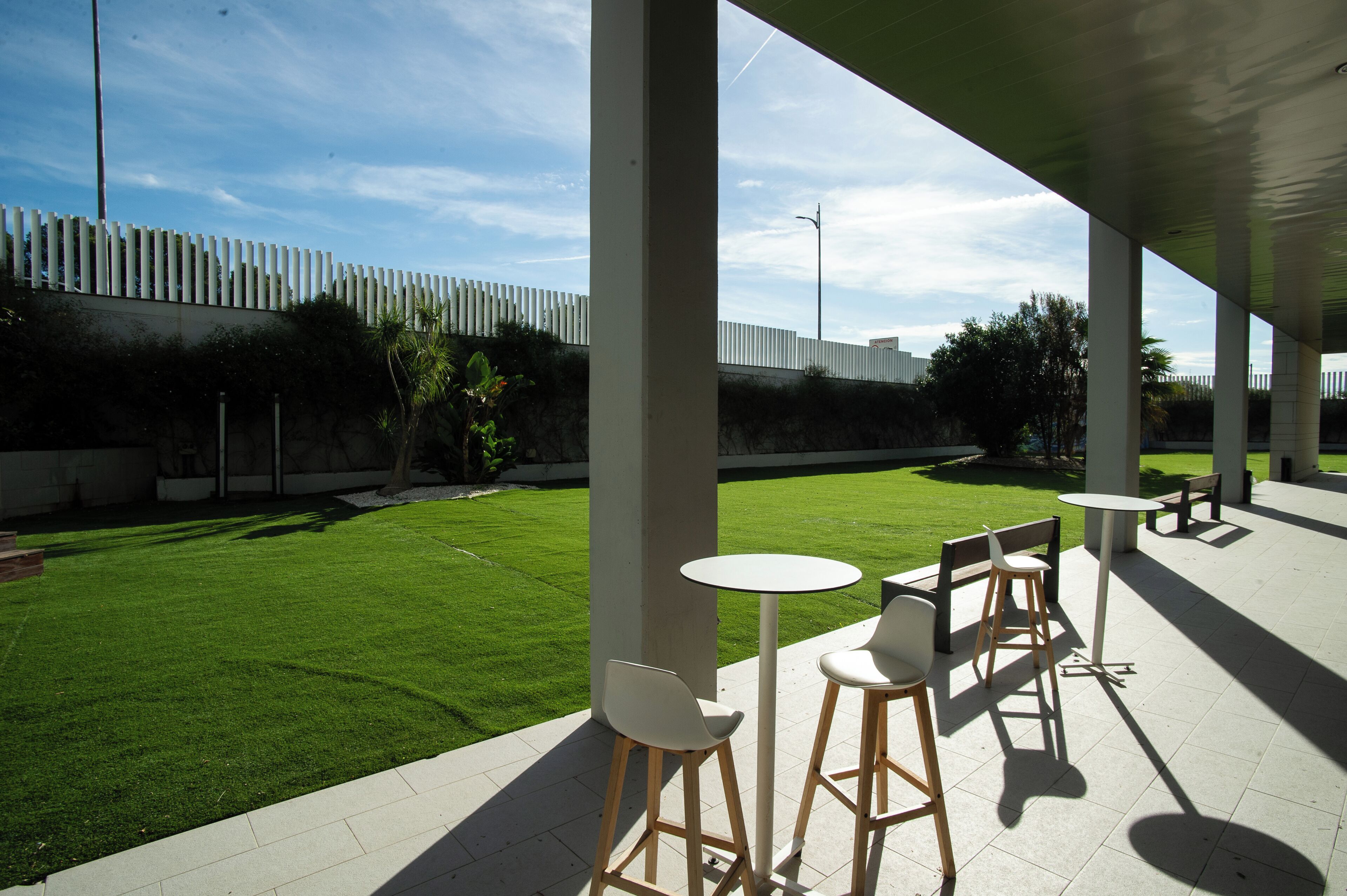 Un espace extérieur aménagé en terrasse, avec une pelouse bien entretenue, des tables et des chaises blanches, et une clôture blanche en arrière-plan.