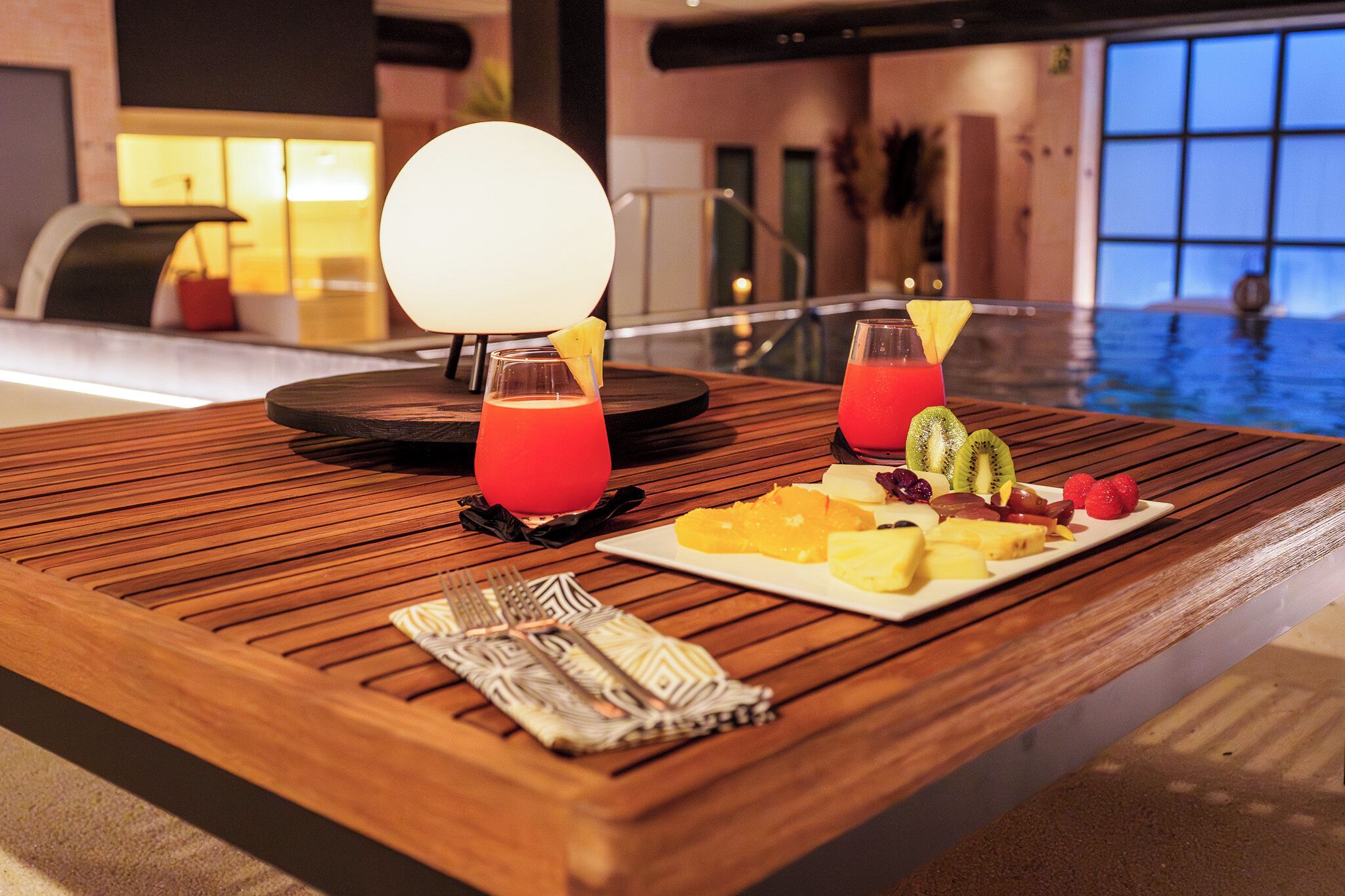 A wooden table with a plate of fruit and two drinks, a lamp, and a view of an indoor pool.