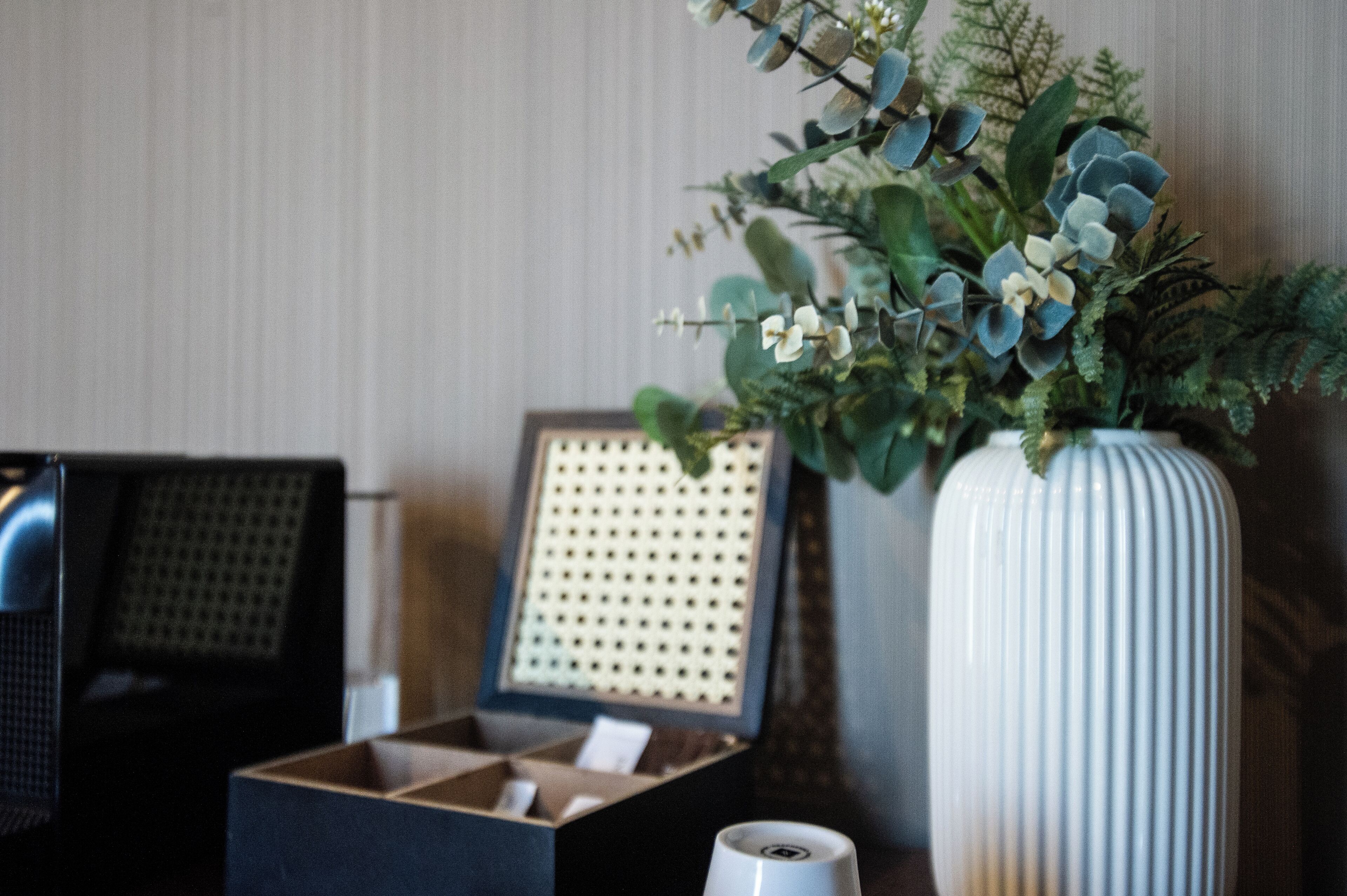 A room with a large white vase with green foliage, a framed picture, and a black box with a white object inside.