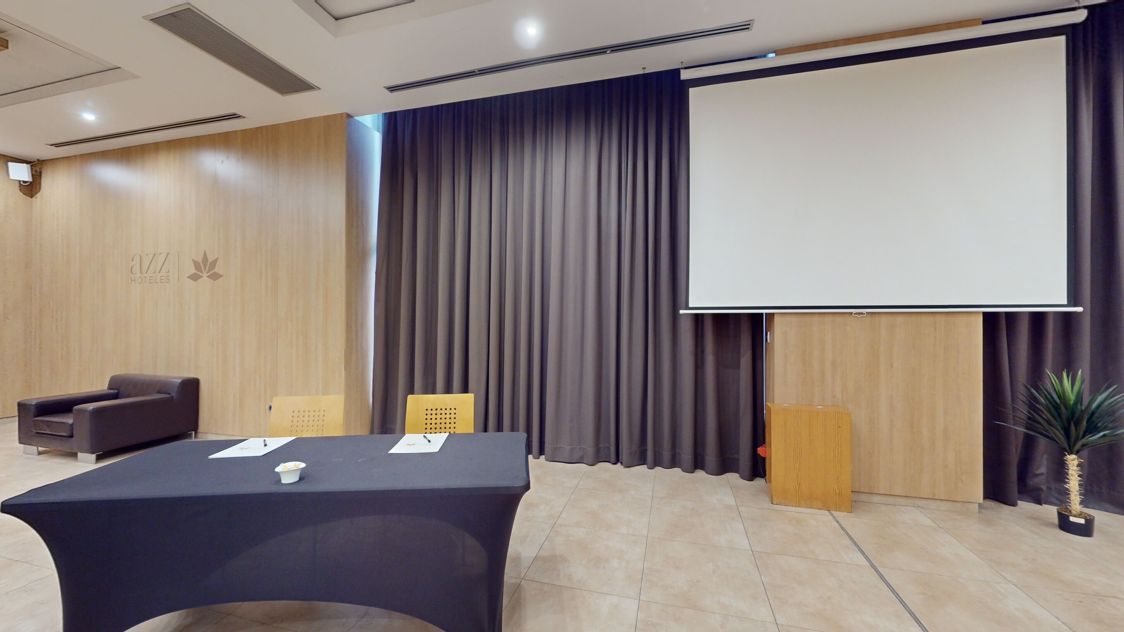 Un salón de conferencias con una mesa, sillas, una pantalla de proyector y una planta en maceta.