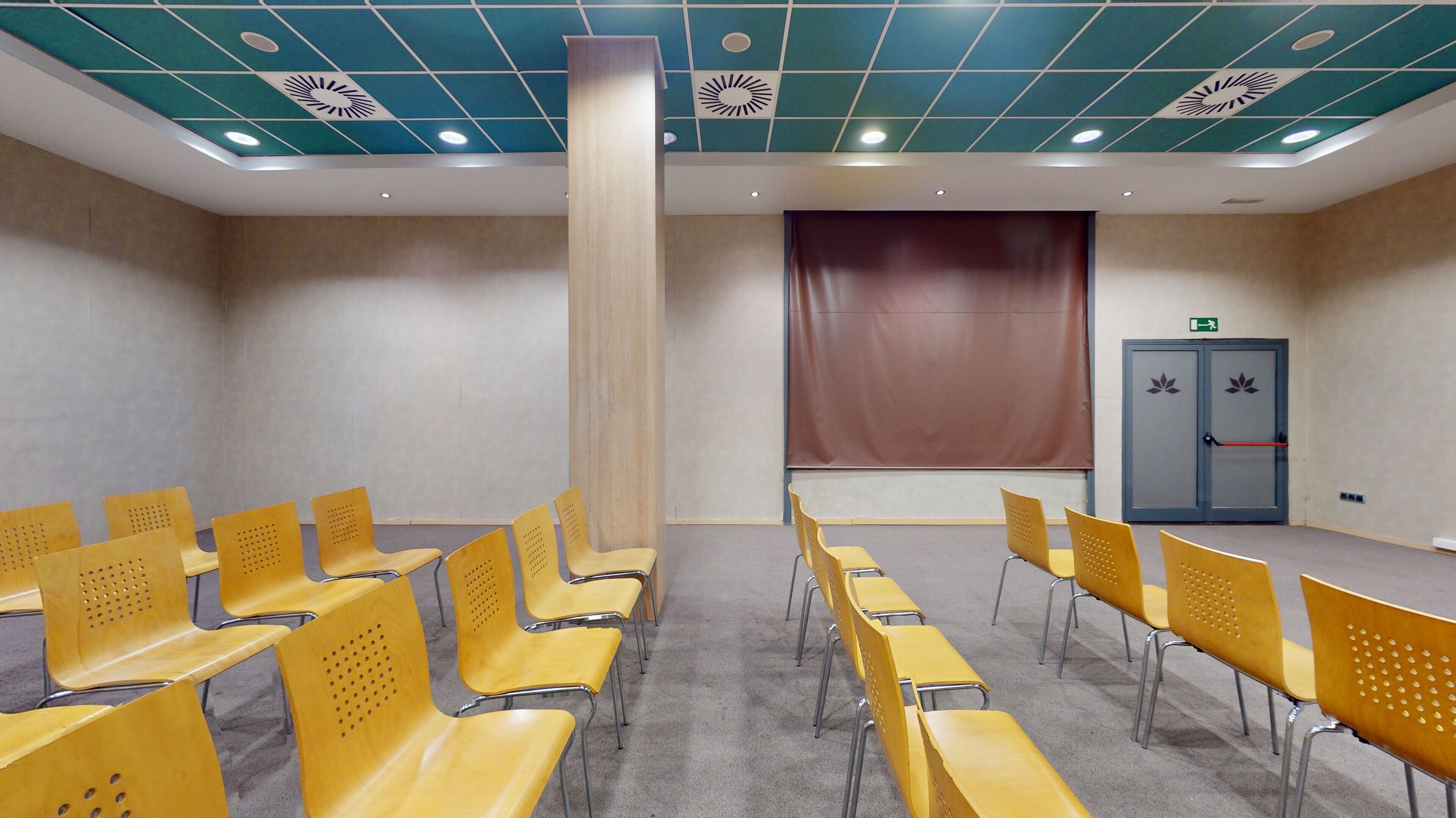 Une salle de conférence avec des chaises jaunes disposées en rangées, un grand rideau au fond et une porte avec une poignée et un bouton rouge sur le côté droit.