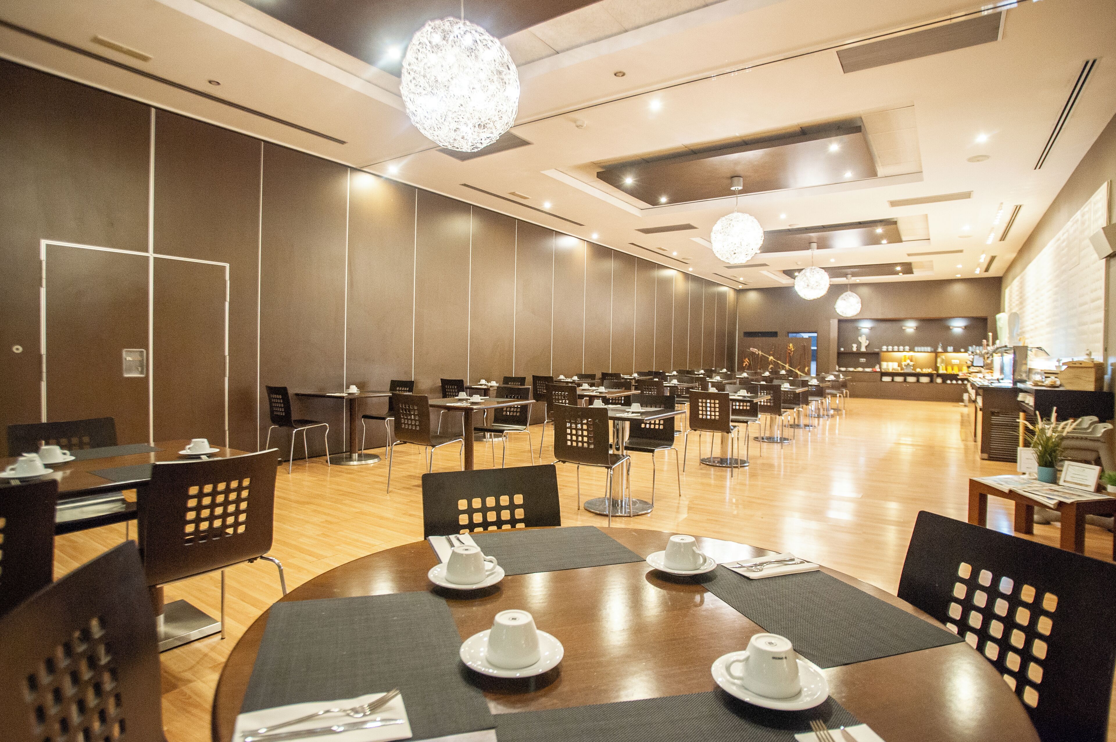 A spacious hotel dining area with multiple tables and chairs, a modern chandelier, and a well-lit interior.