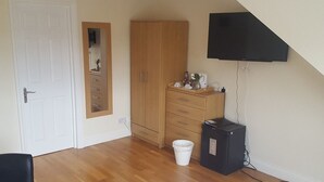 A room with a wooden wardrobe, a small cabinet with drawers, a black TV mounted on the wall, a white trash bin, and a black electrical box. - Kathleen House (Enfield)