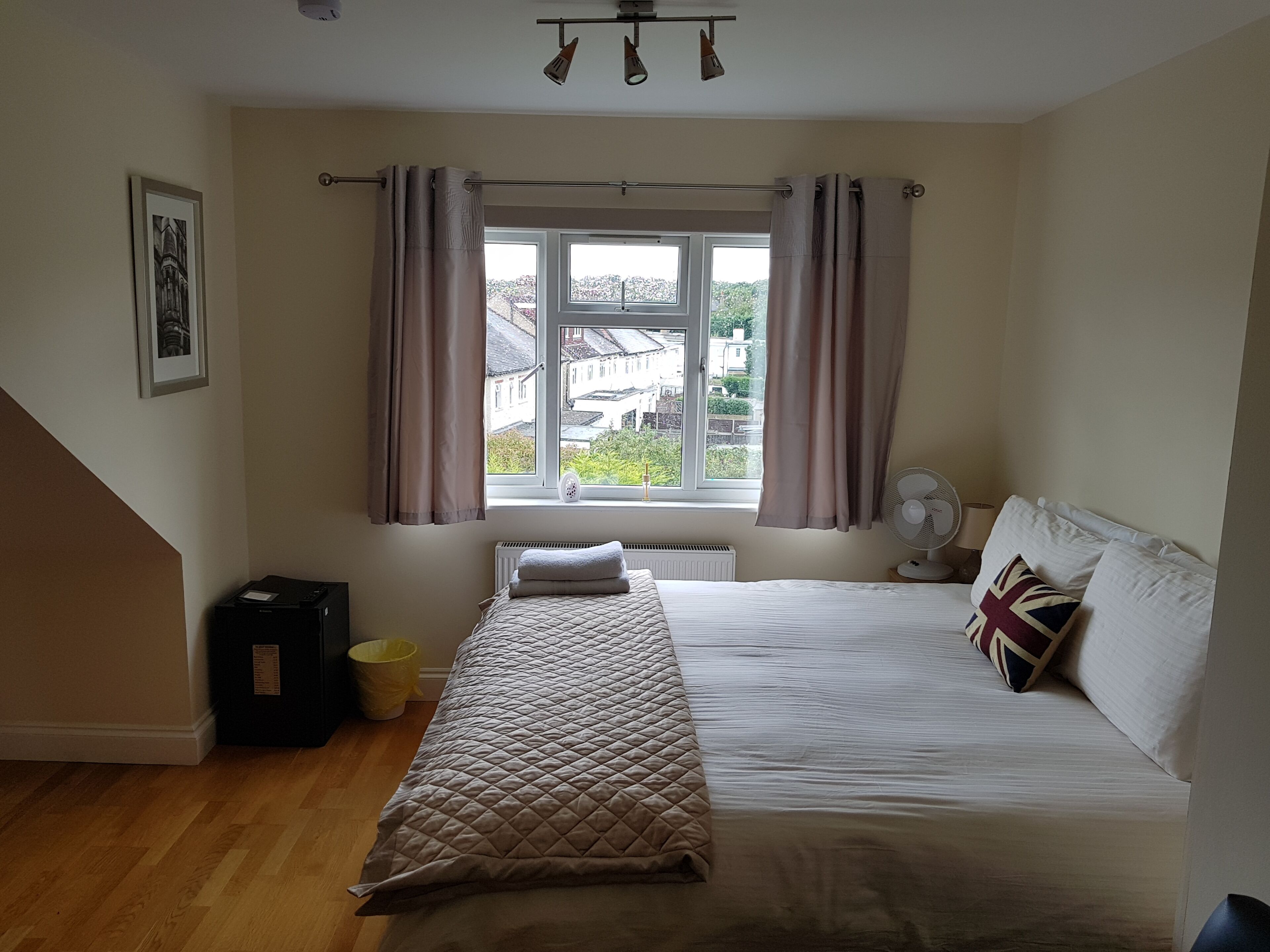 A bedroom with a bed, a window with curtains, a radiator, a fan, and a picture on the wall.
