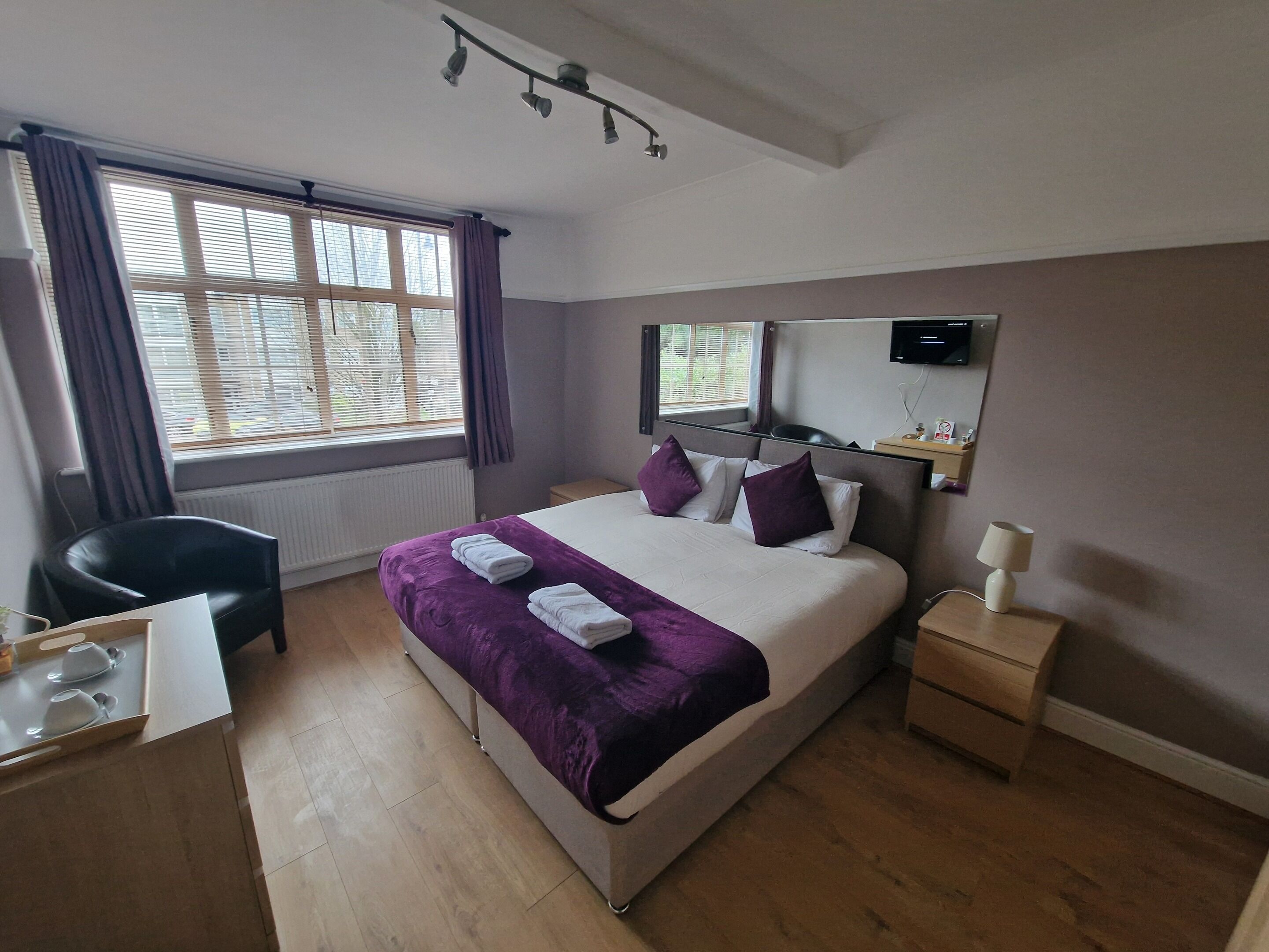A hotel room with a bed, a desk, a chair, a bedside table, a mirror, and a window with curtains.