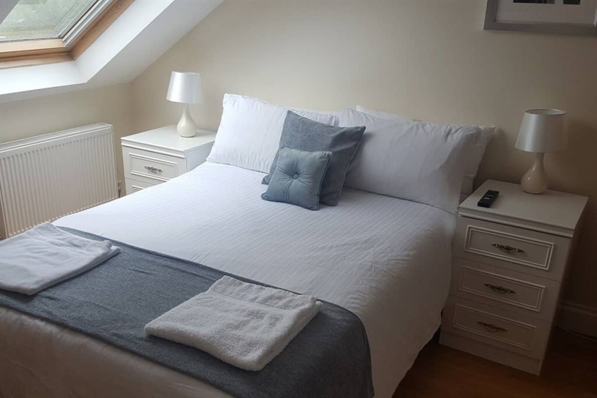 Ein ordentlich bezogenes Bett mit weißen Laken und einer grauen Decke, ein Nachttisch mit Lampe sowie ein weiterer Nachttisch mit Telefon.