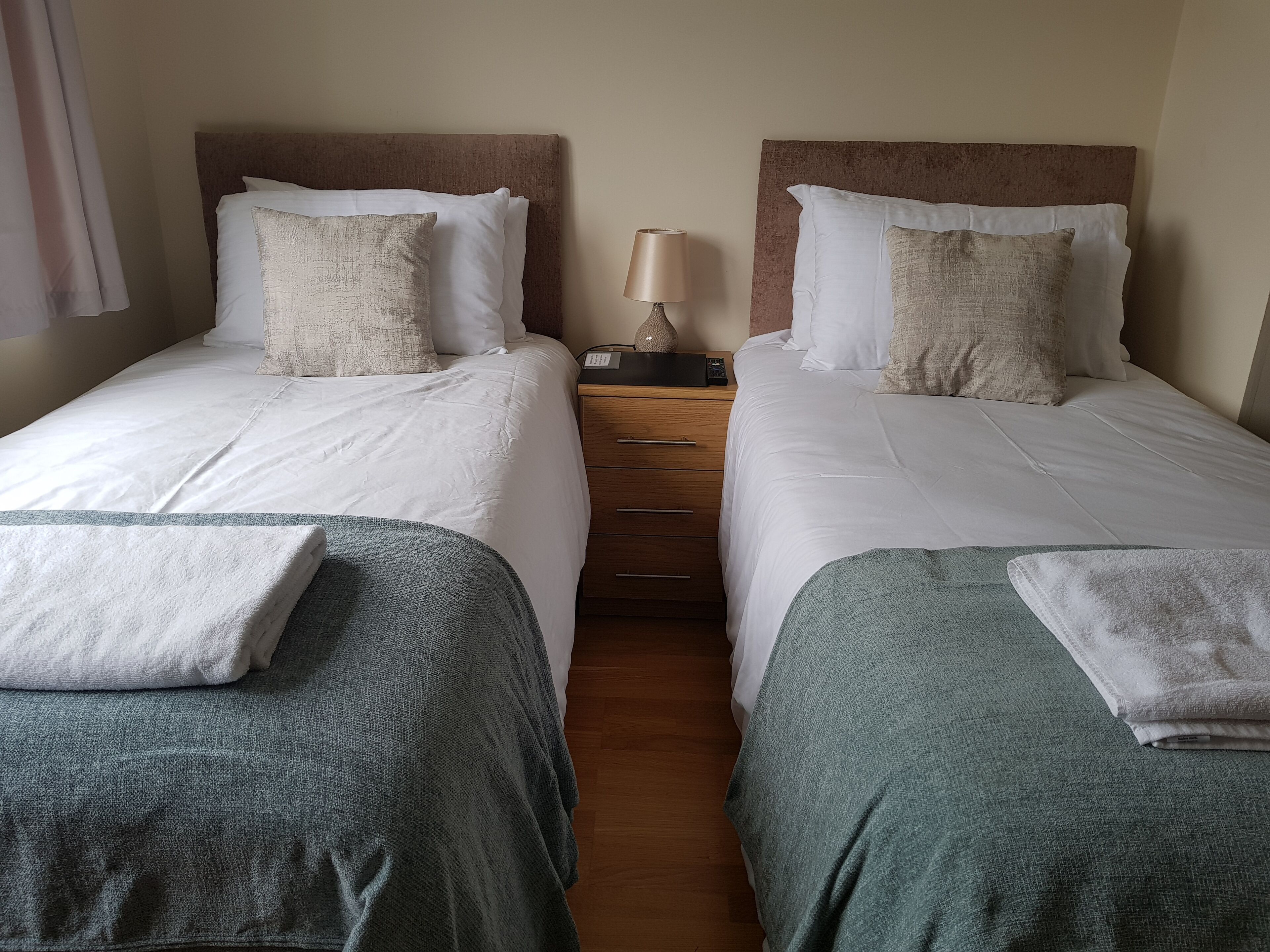 A double room with two beds, each with white linens and a grey throw blanket. There is a wooden nightstand with a lamp between the beds.