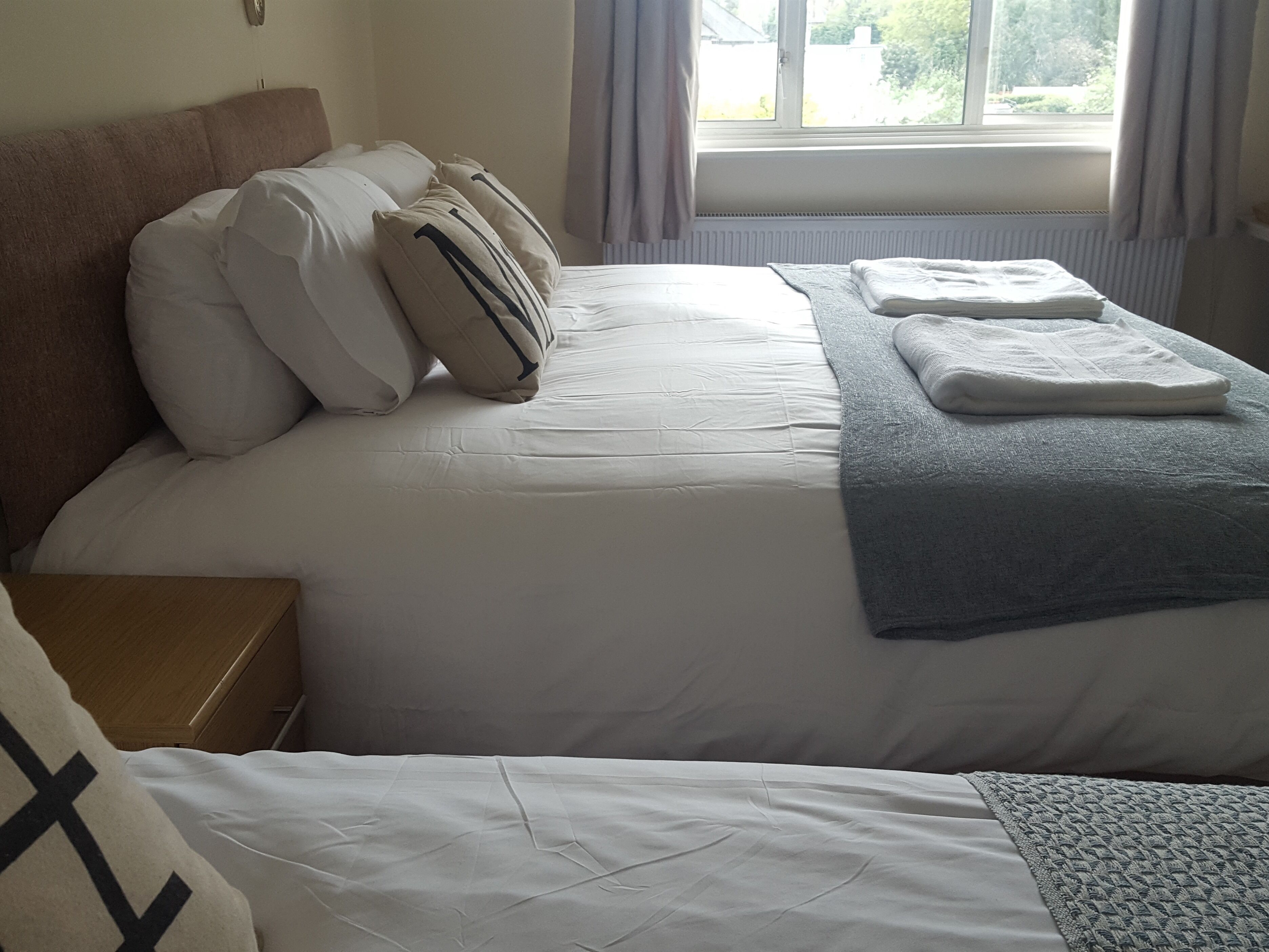Un letto rifatto con lenzuola bianche e una coperta grigia, un comodino di legno e una finestra con tende.