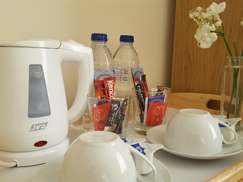 A table with a white electric kettle, two bottles of water, a glass with a red packet, a glass with a blue packet, a white cup, and a saucer.