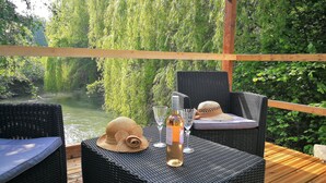 A wicker outdoor seating area with a bottle and glasses on a table, overlooking a river and greenery.