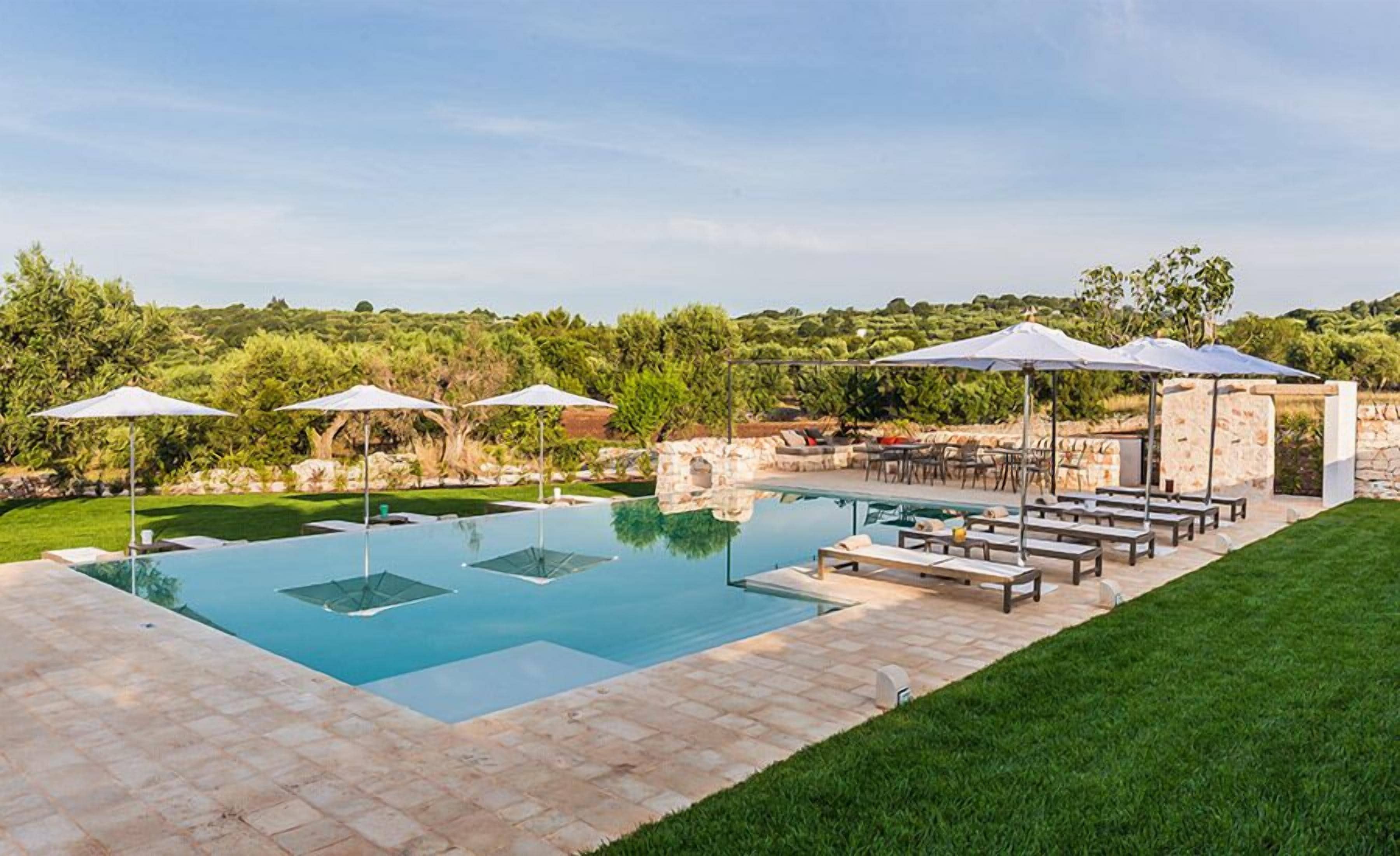 Una piscina rectangular con agua azul cristalina, rodeada por un patio de piedra. Hay múltiples sillas de descanso y sombrillas que brindan sombra. La zona está ajardinada con césped verde y árboles al fondo.