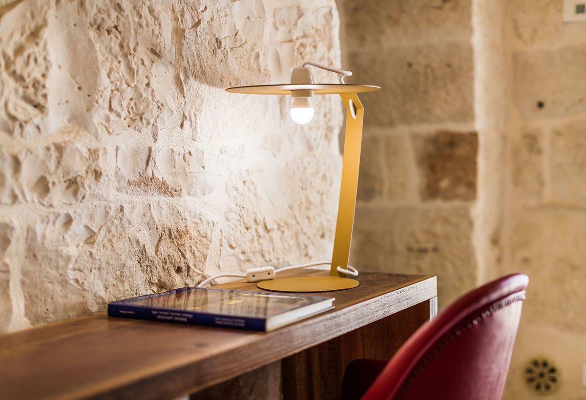 Una lámpara sobre una mesa de madera, con un libro y una silla roja en una habitación con paredes de piedra.