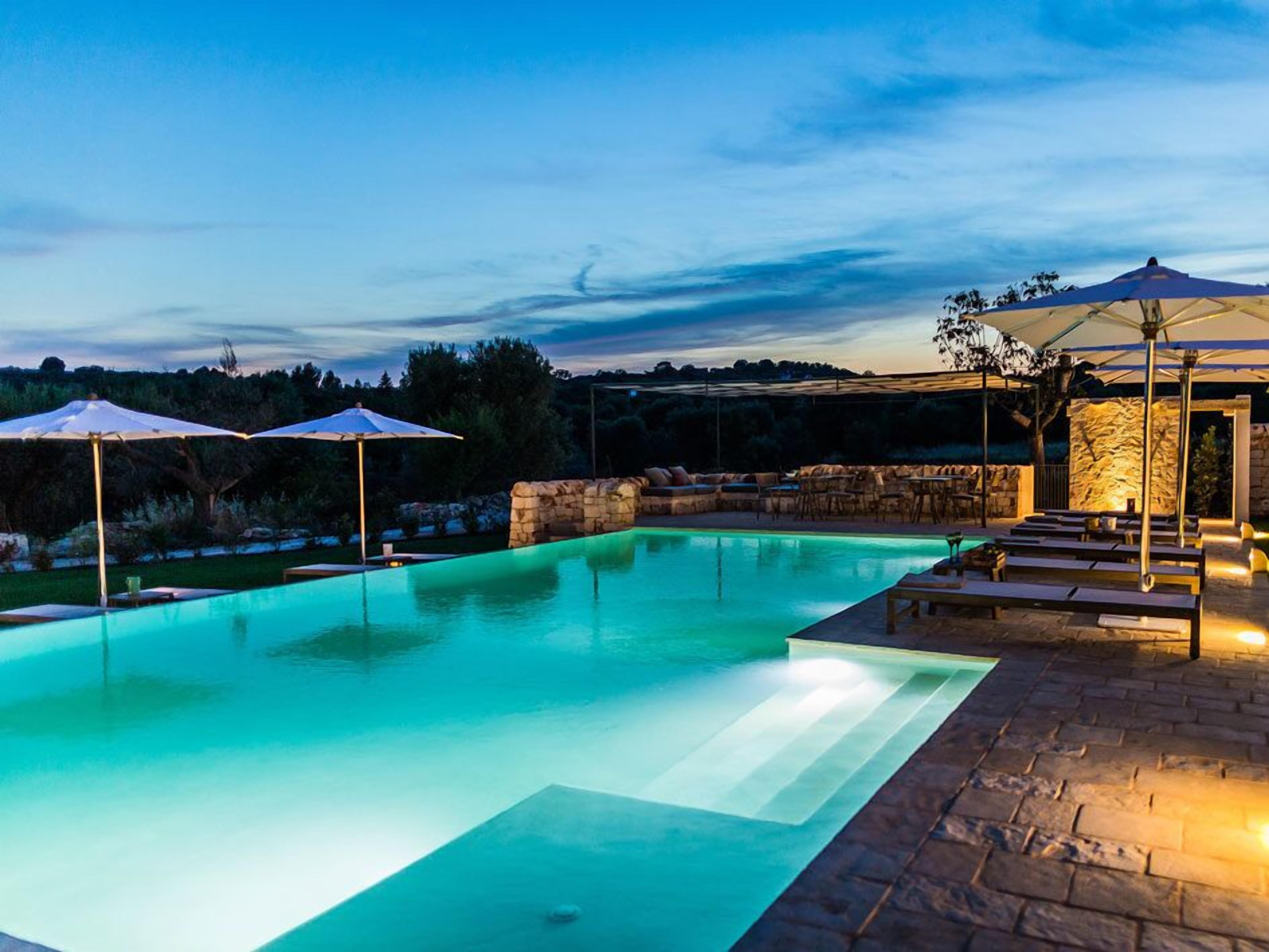 Una piscina con muretto in pietra e zona relax, circondata da ombrelloni e una struttura in pietra.