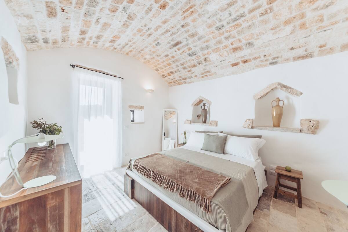 A bedroom with a stone ceiling, a wooden desk, a bed with a beige blanket, and a small stool.