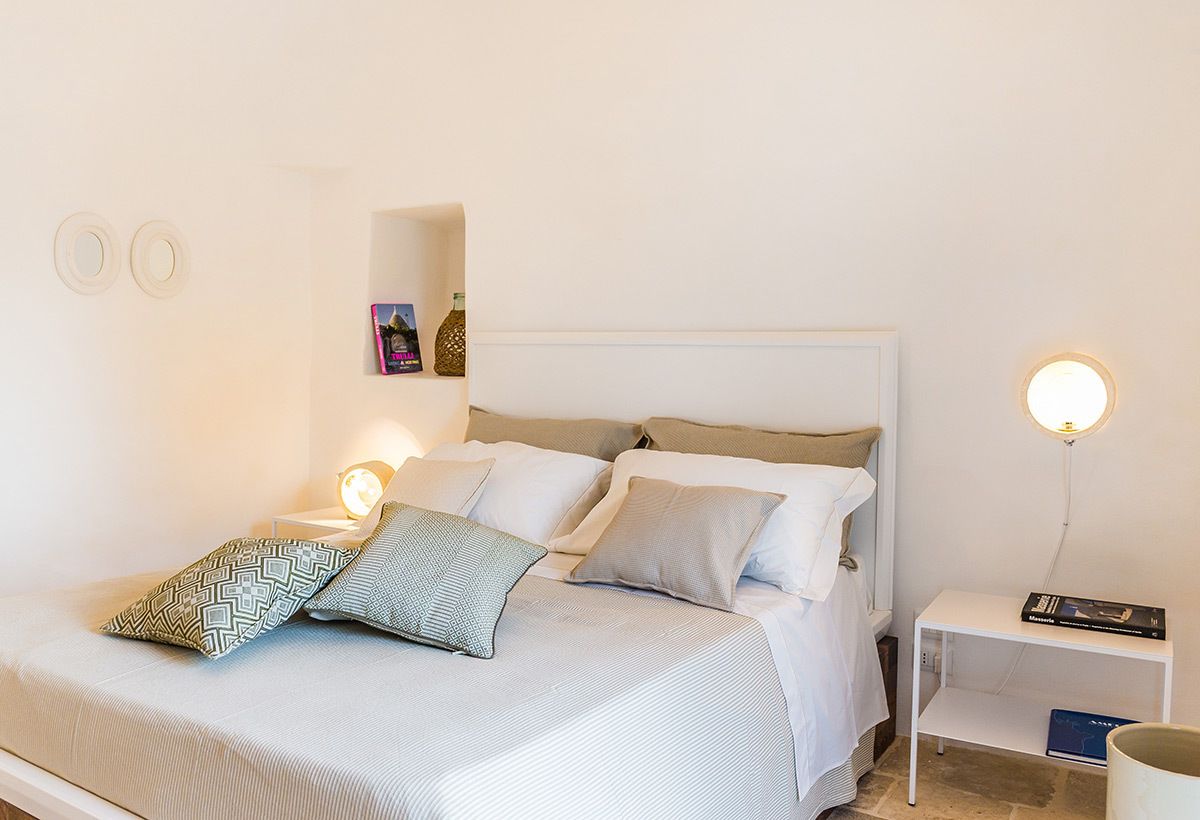 Una cama bien hecha con almohadones estampados, una mesita de noche con lámpara y una pequeña estantería con libros.