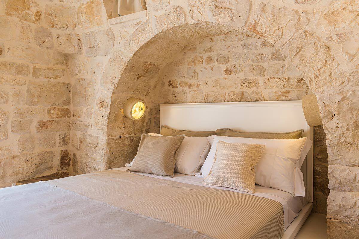 A bedroom with a stone wall, a bed with beige bedding, and a wall-mounted light.
