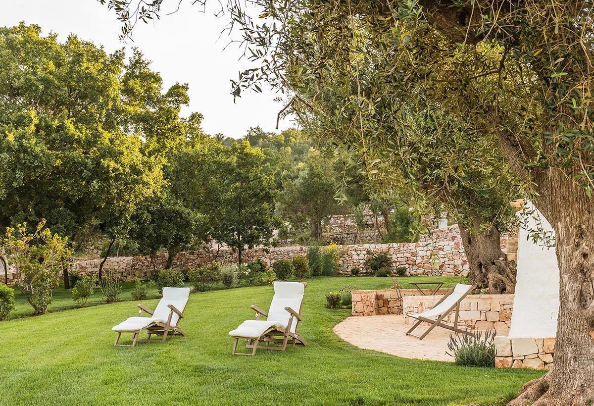 Een tuin met drie witte lounge fauteuils, een stenen muur en weelderige begroeiing.