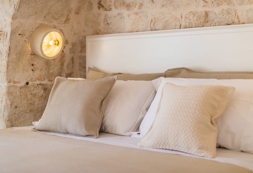 Cama com roupa de cama branca e almofadas bege em quarto com parede de pedra e luminária fixada na parede.