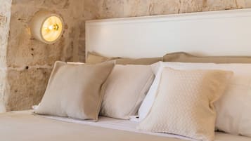 A bed with white bedding and beige pillows in a room with a stone wall and a wall-mounted light fixture.