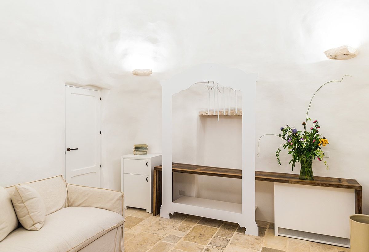 A room with a beige sofa, a white cabinet, a wooden shelf, and a vase with flowers.